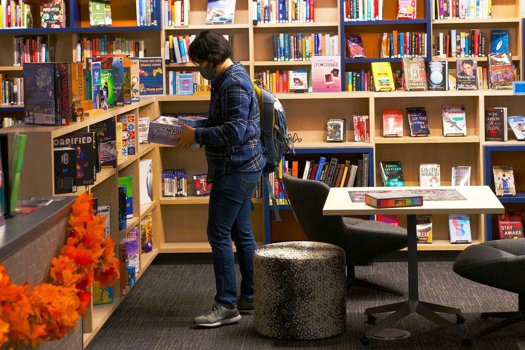 Student with mask holding stack of board games in ISR library